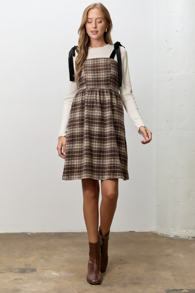 woman wearing a brown plaid dress with black ribbon straps over a white long sleeve top and brown ankle boots