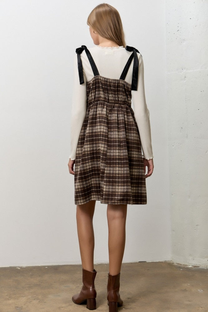 woman wearing a brown plaid dress with black straps over a cream sweater and brown ankle boots, seen from the back