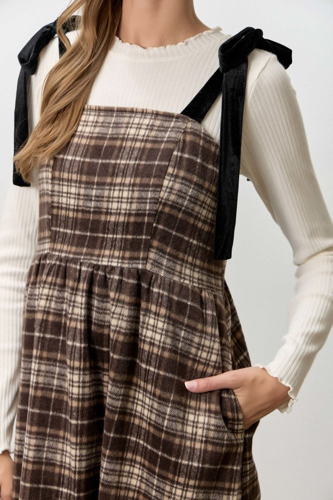 woman wearing a brown plaid overall dress with black ribbon straps over a cream ribbed long sleeve top
