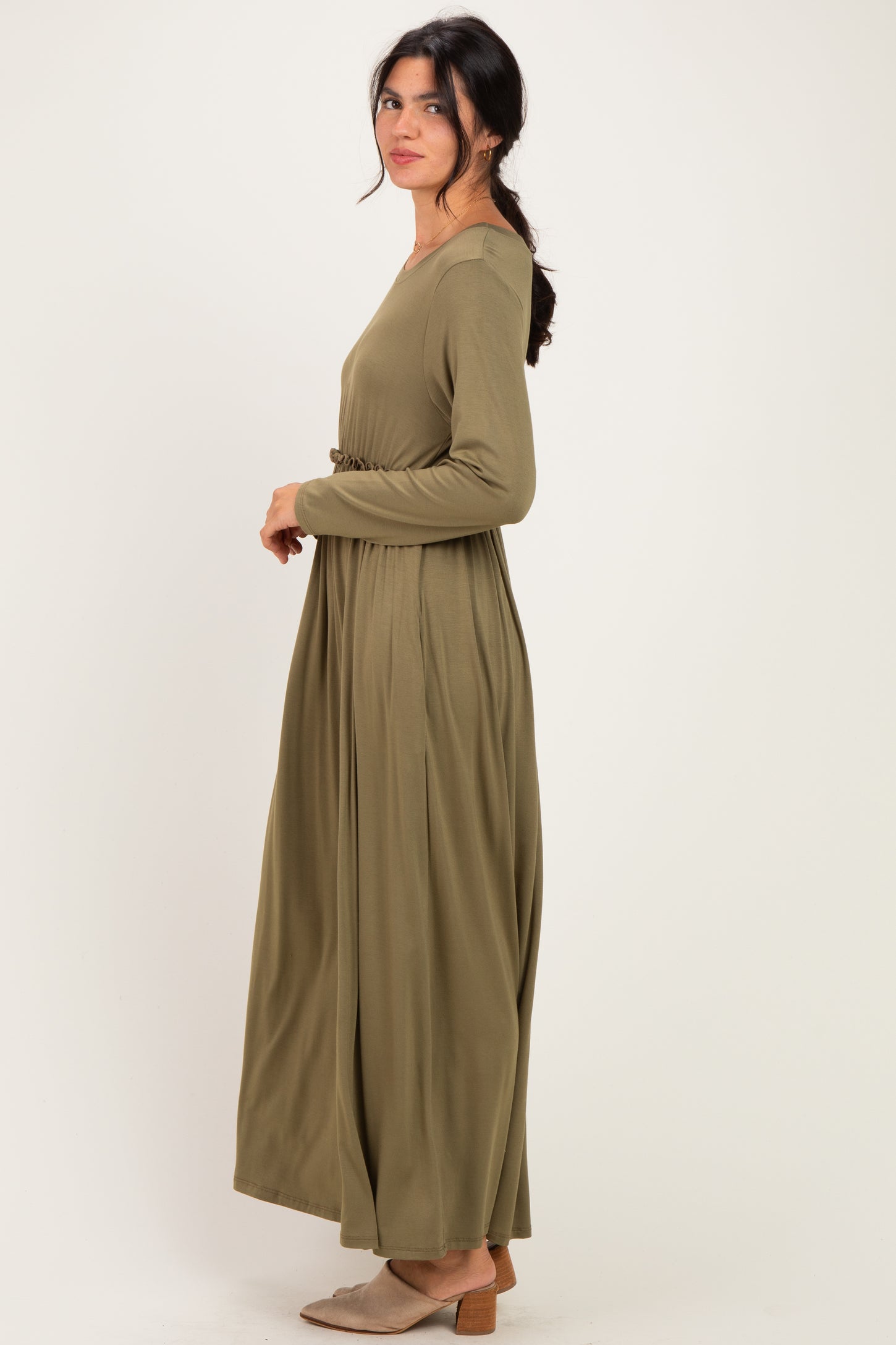 woman wearing an olive green long sleeve maxi dress with a waist tie, standing sideways against a light background