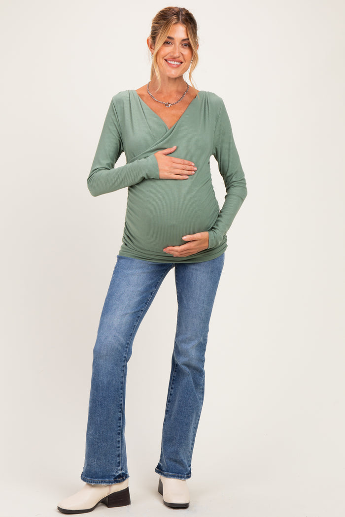 woman wearing a green long sleeve maternity top and blue flared jeans, holding her baby bump