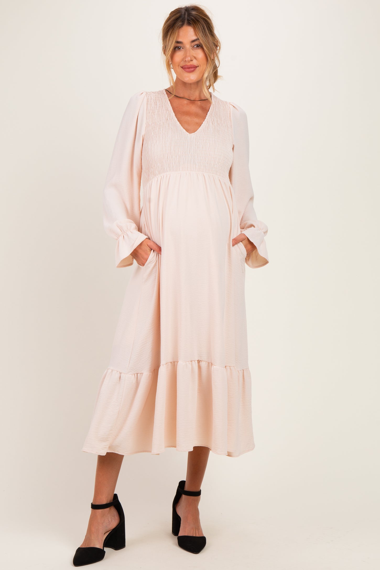 woman wearing a blush pink maternity dress with a smocked bodice and long sleeves, featuring a tiered skirt and pockets