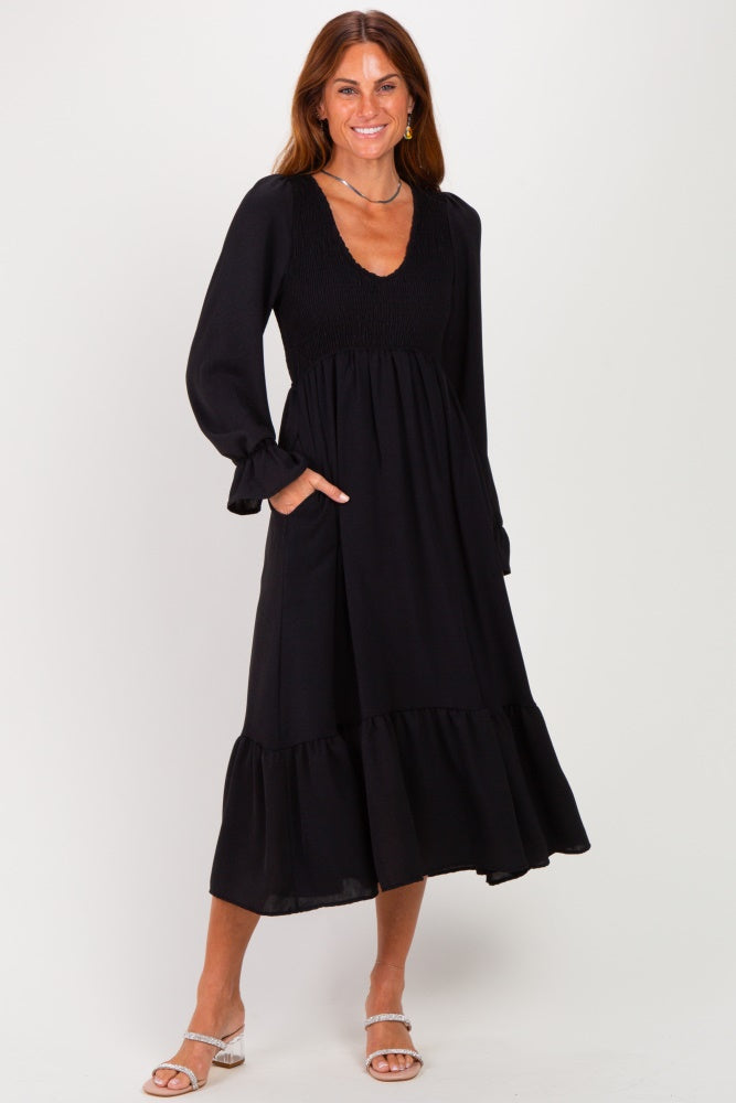 woman wearing a black long sleeve midi dress with smocked bodice and tiered hem, posing with hands in pockets