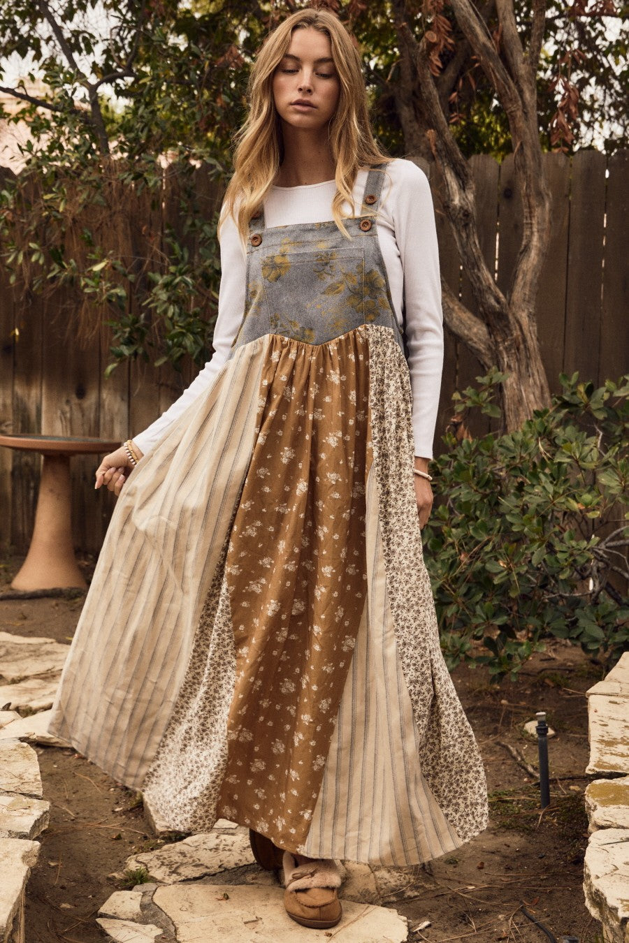 woman wearing a bohemian-style long patchwork dress with suspenders, featuring floral and striped patterns in earth tones