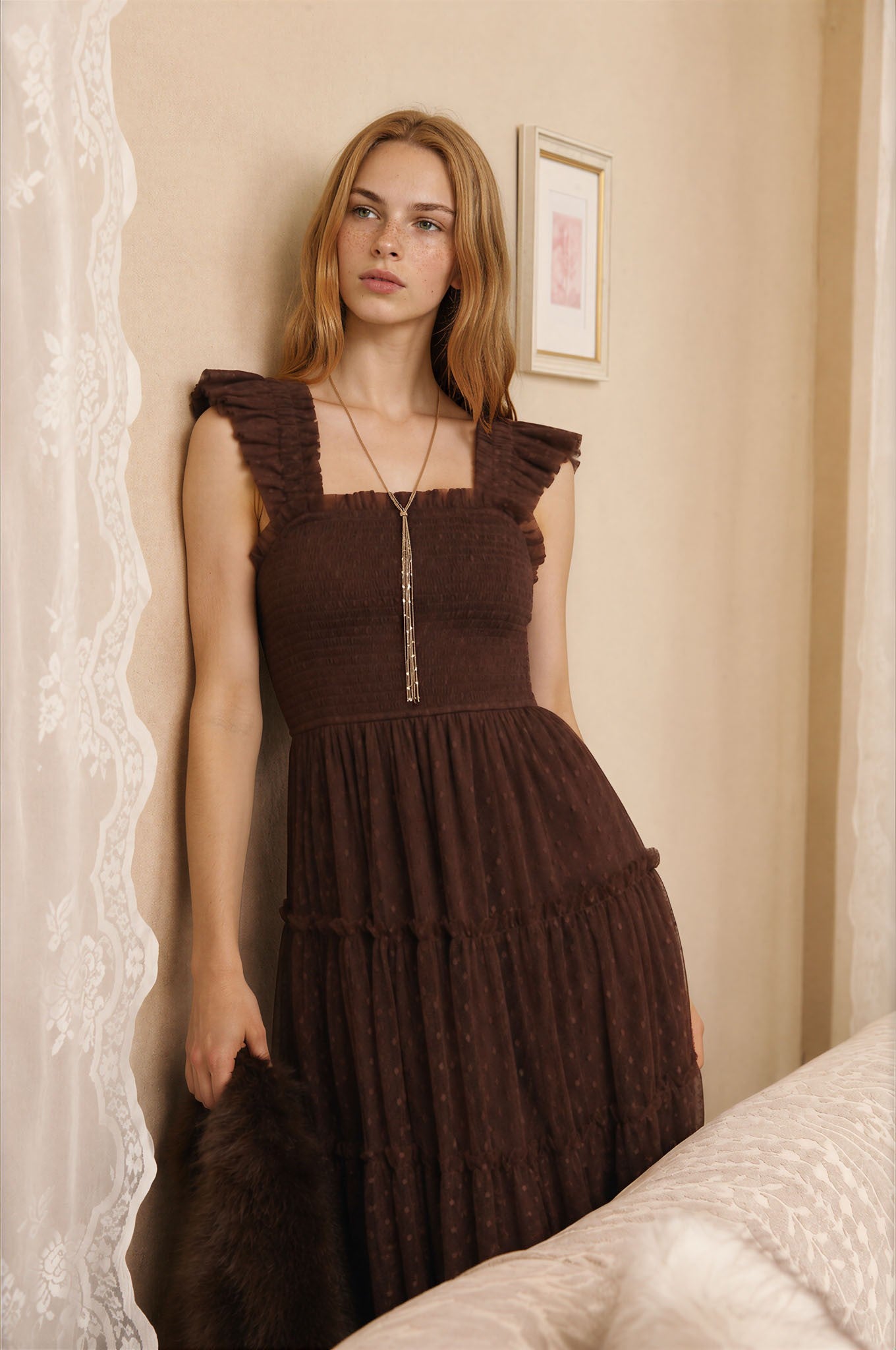 woman wearing a brown tiered dress with ruffled straps and a fitted bodice, standing by a wall with lace curtains