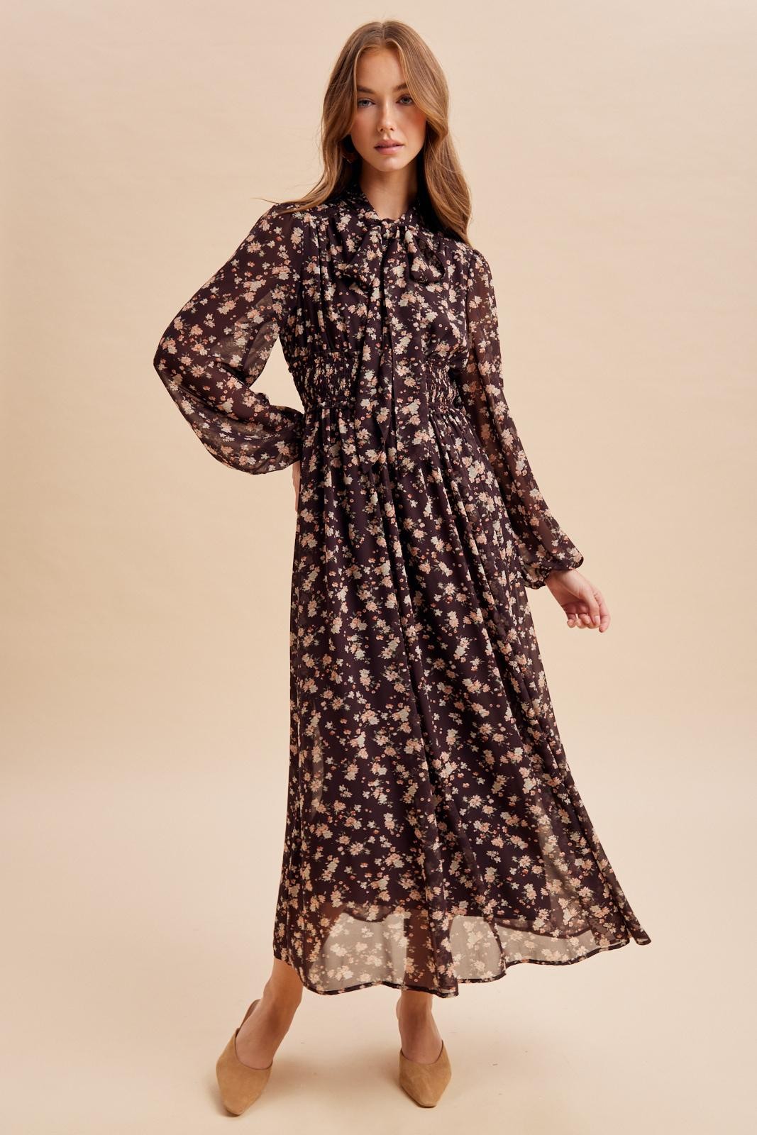 woman wearing a long sleeve black floral maxi dress with a cinched waist and ruffled collar, posing against a light background