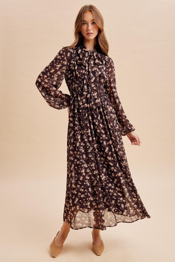 woman wearing a long sleeve black floral maxi dress with a cinched waist and ruffled collar, posing against a light background