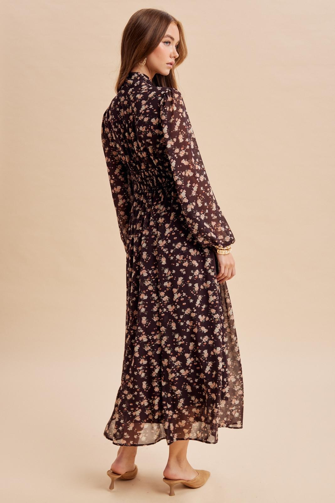 woman wearing a black floral maxi dress with long sleeves and back ruching, styled with nude heels