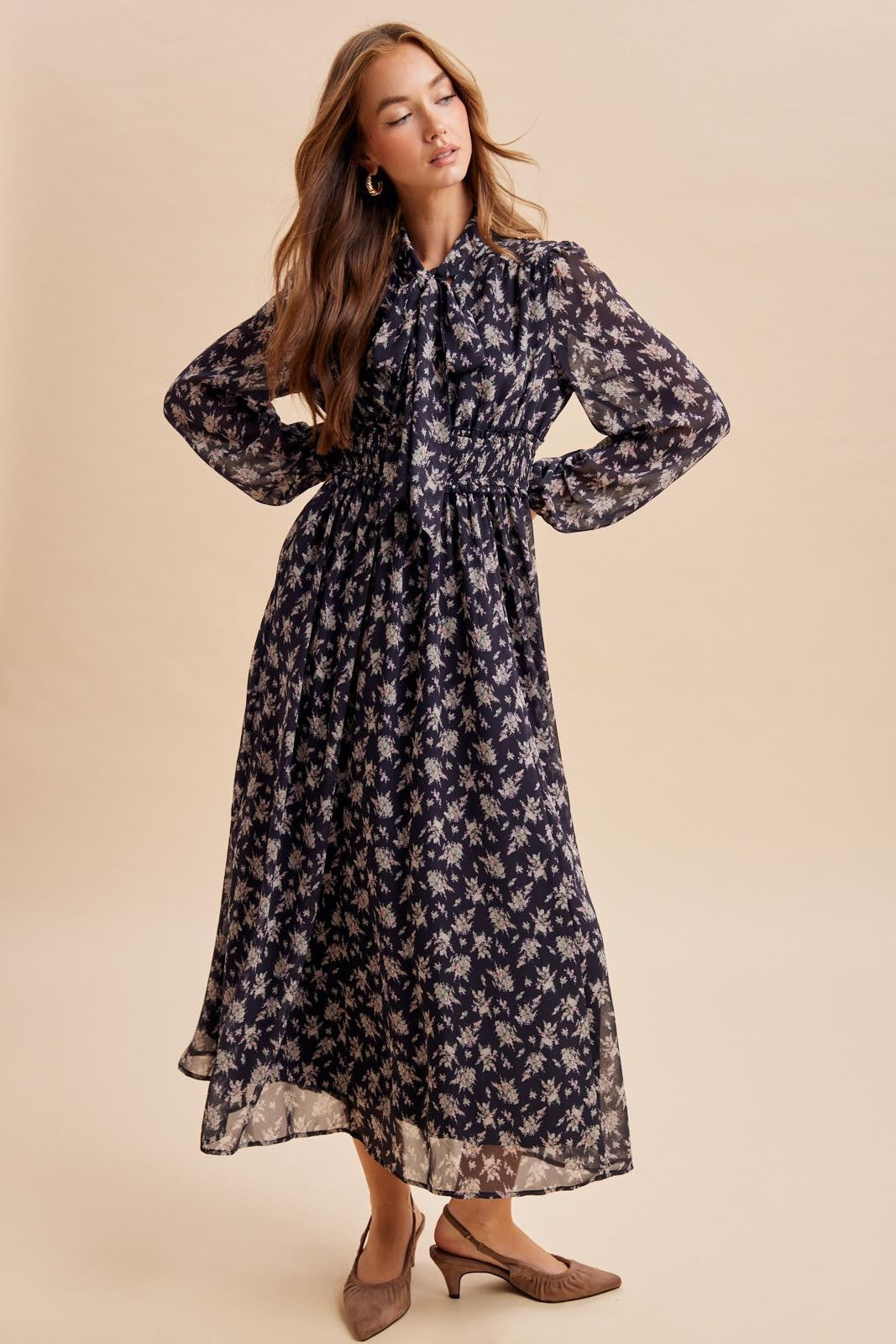 woman wearing a navy blue floral maxi dress with long sleeves and a tie waist detail, styled with brown heeled mules