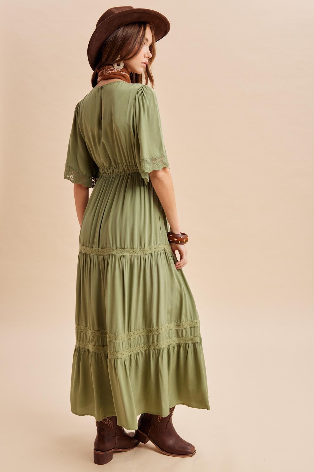 woman wearing a green maxi dress with short sleeves and lace details, accessorized with a brown hat and boots