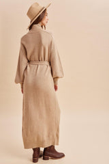 woman wearing a beige long sleeve cardigan dress with a belted waist and brown ankle boots, facing away from the camera