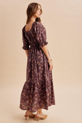 woman wearing a purple floral maxi dress with puff sleeves and a tiered skirt, styled with tan mules and a gold bracelet