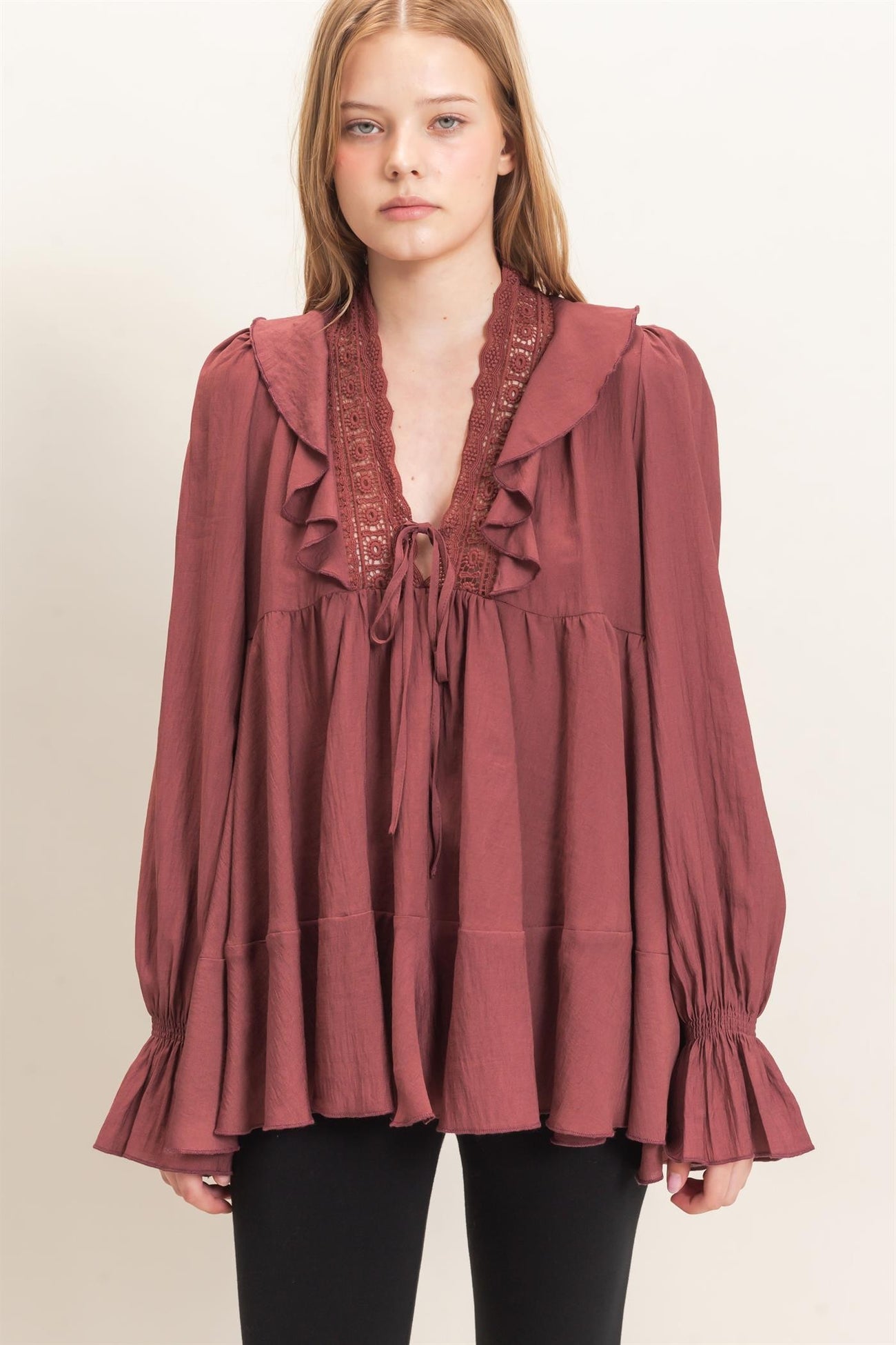 woman wearing a burgundy long sleeve top with ruffle details and a tie neckline, paired with black pants
