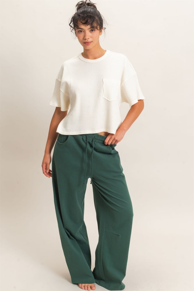 woman wearing a white ribbed short sleeve top and green wide leg pants, standing barefoot against a neutral background
