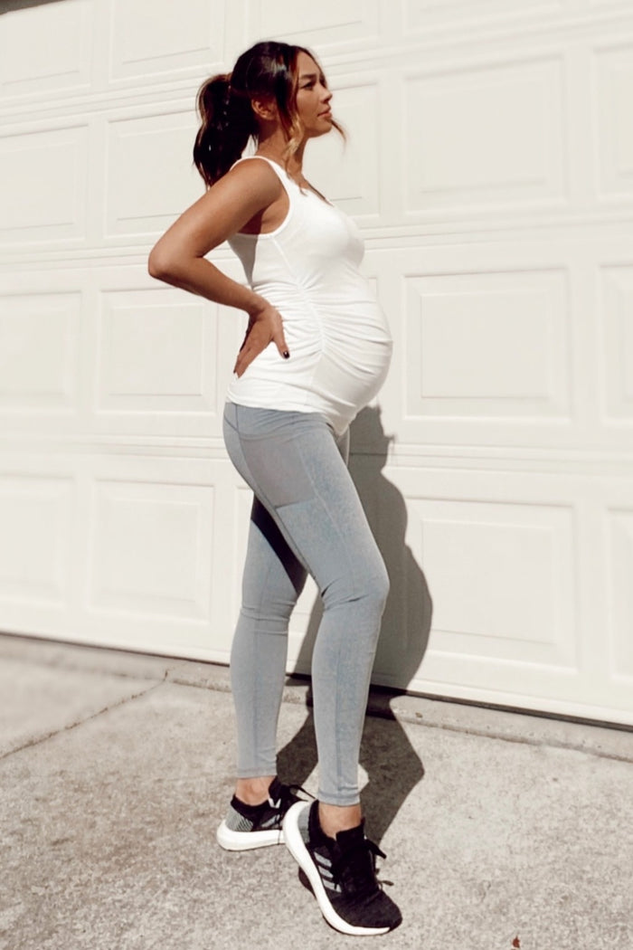 woman wearing a white maternity tank top and light blue maternity leggings, showcasing a visible baby bump
