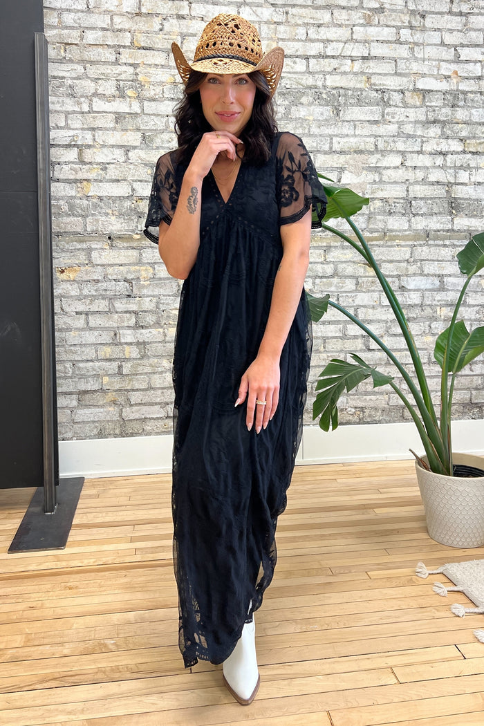 woman wearing a black maxi dress with lace details and short sleeves, accessorized with a straw cowboy hat and white ankle boots