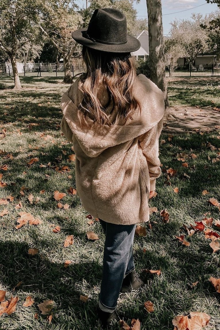 woman wearing a tan fuzzy hooded jacket, black hat, and dark jeans, standing in a park surrounded by autumn leaves