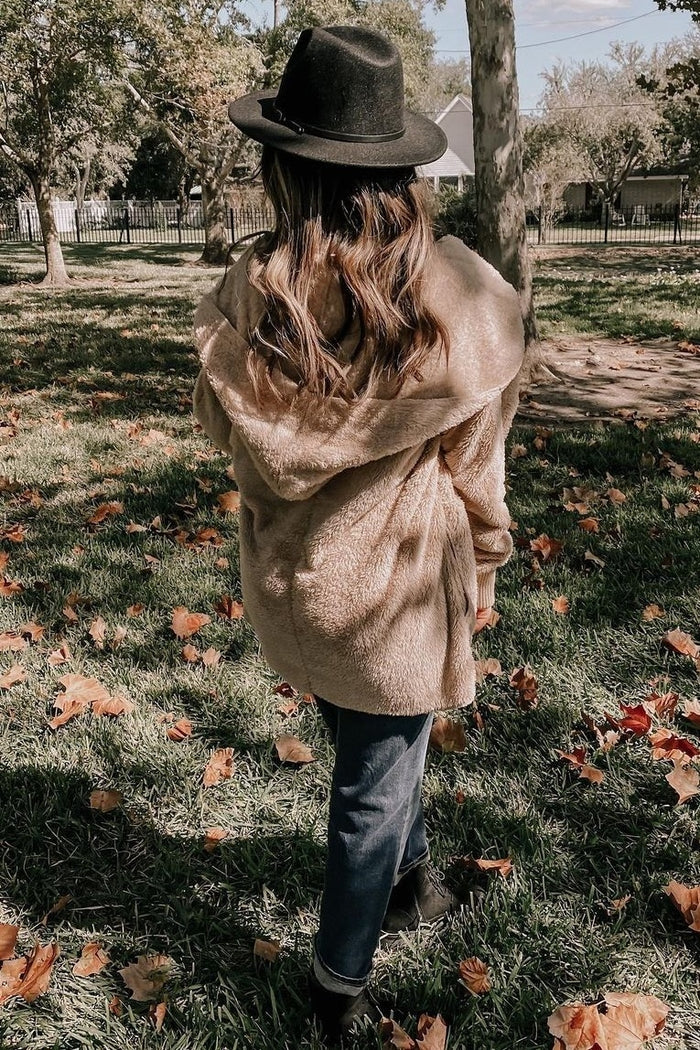 woman wearing a tan fuzzy hooded jacket, black hat, and dark jeans, standing in a park surrounded by autumn leaves
