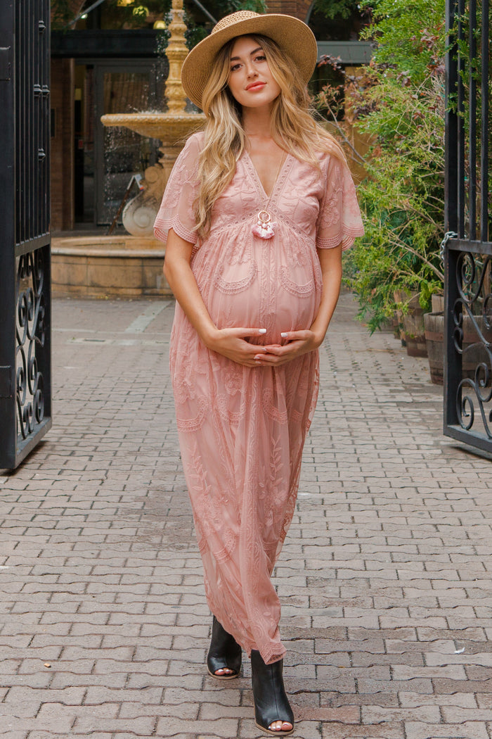 woman wearing a pink lace maternity dress sitting in a field and cradling her baby bump while holding flowers
