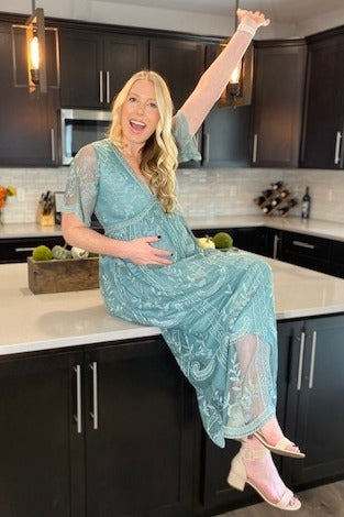 woman wearing a teal maternity dress with lace details and a visible baby bump, sitting on a kitchen counter and smiling