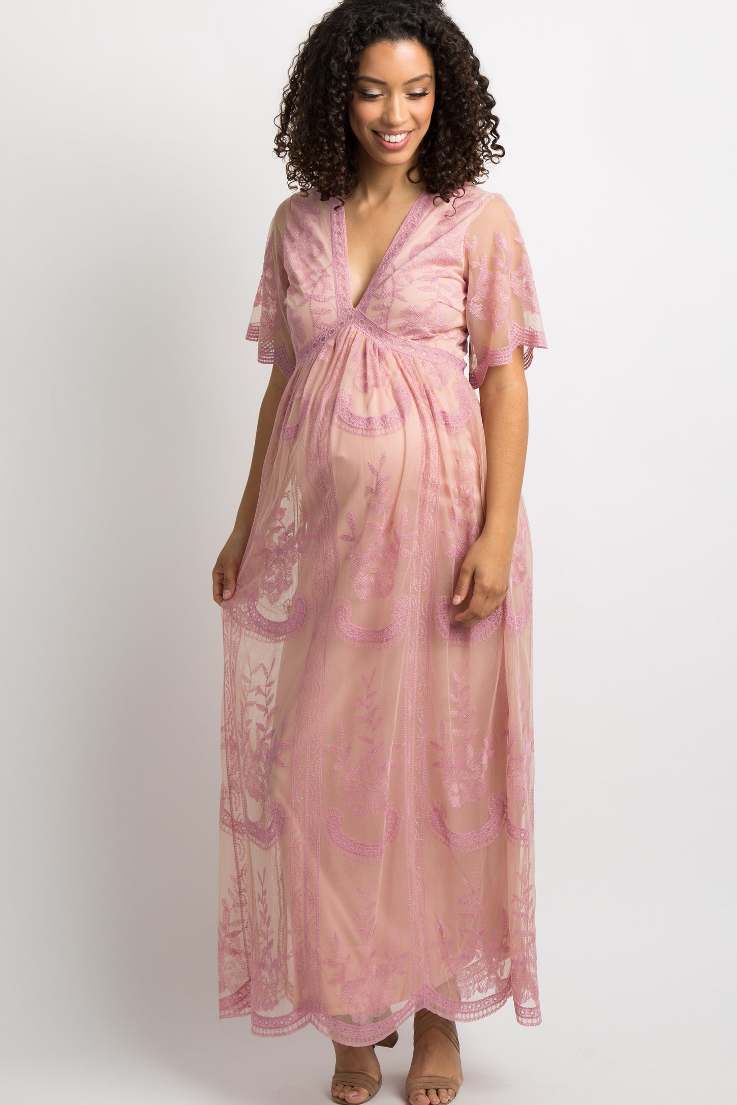 woman wearing a sheer pink maternity dress with lace details, smiling and holding her baby bump beside a man in a gray t-shirt
