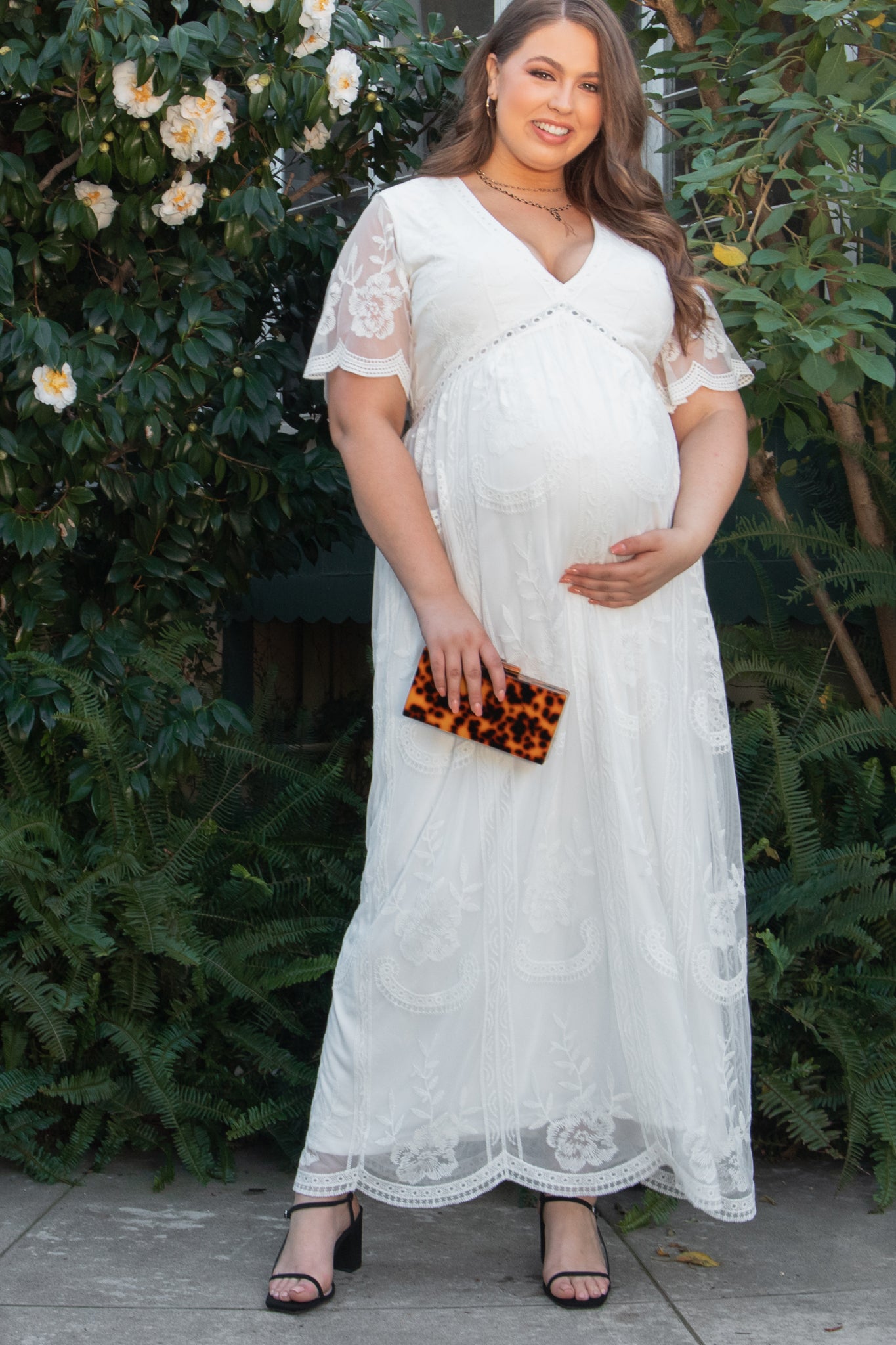 woman wearing a white lace maternity maxi dress and holding her baby bump with a small clutch bag