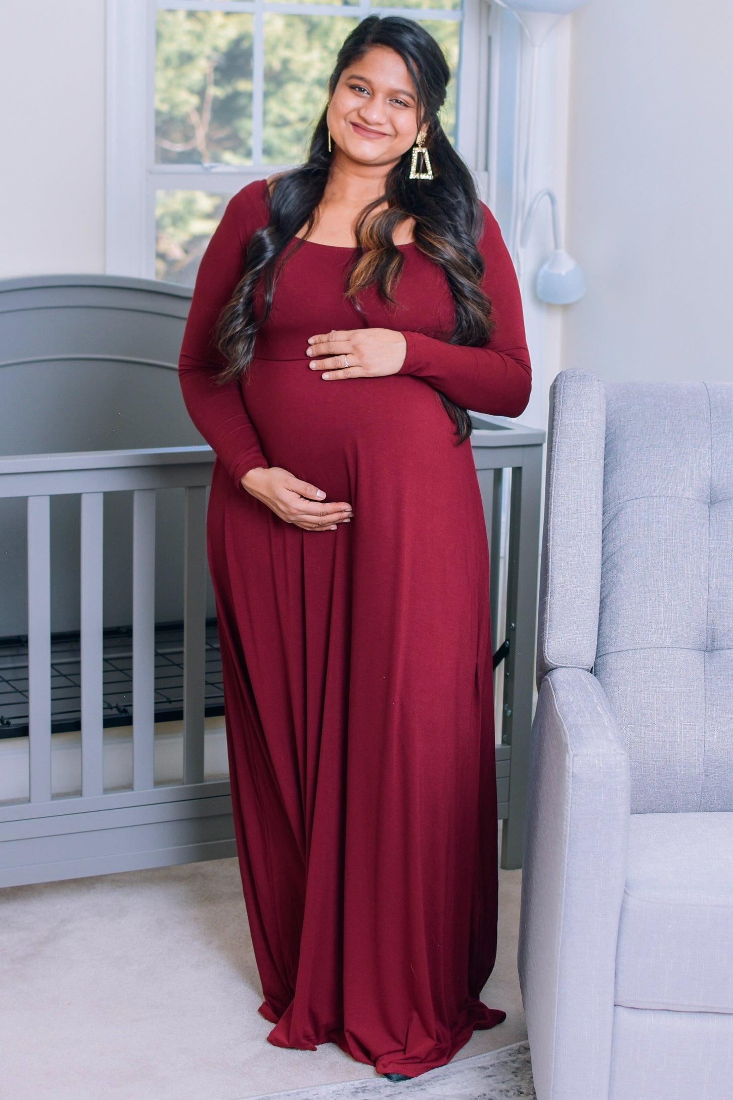 woman wearing a burgundy long sleeve maternity gown and holding her baby bump while standing beside a crib