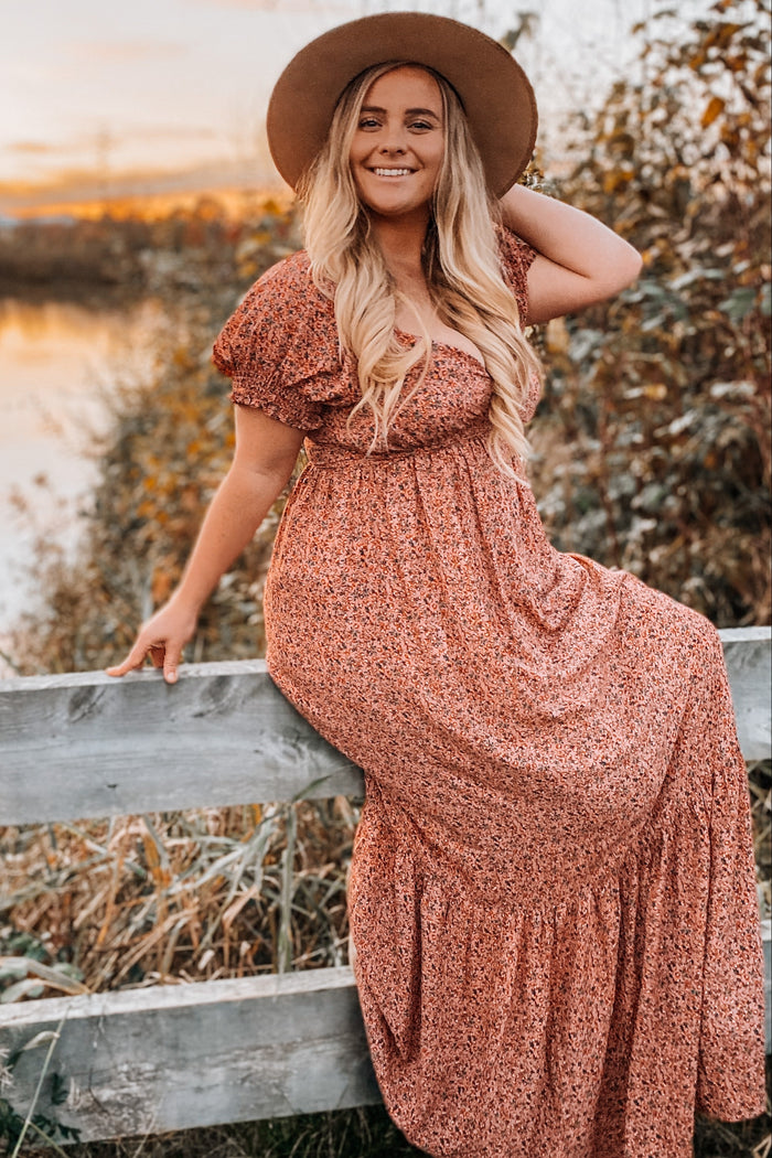 woman wearing a floral print maxi dress with short ruffled sleeves and a wide-brimmed hat, posing on a fence in a natural setting