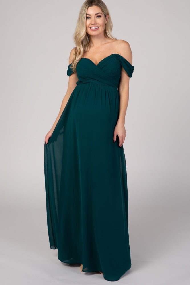 woman wearing a dark green off-the-shoulder maternity gown with a flowing skirt and ruched bodice, showcasing her baby bump