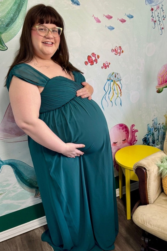 woman wearing a teal maternity gown with a sweetheart neckline and cap sleeves, gently cradling her baby bump