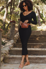 woman wearing a black ribbed midi dress and sandals, holding a brown waist bag, standing on outdoor stairs in a park