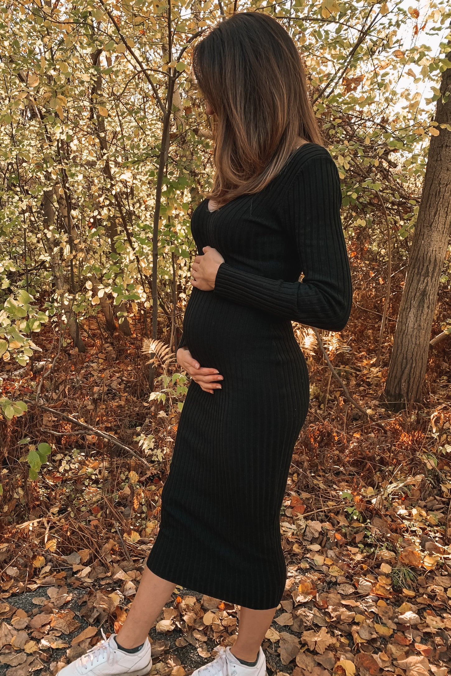woman wearing a black ribbed maternity dress and holding her baby bump, standing in a forest with autumn leaves