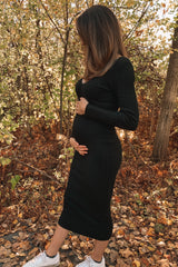 woman wearing a black ribbed maternity dress and holding her baby bump, standing in a forest with autumn leaves