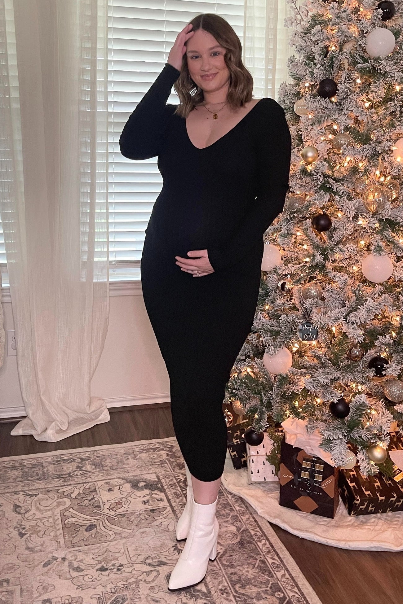 woman wearing a black ribbed maternity dress and holding her baby bump in front of a decorated Christmas tree