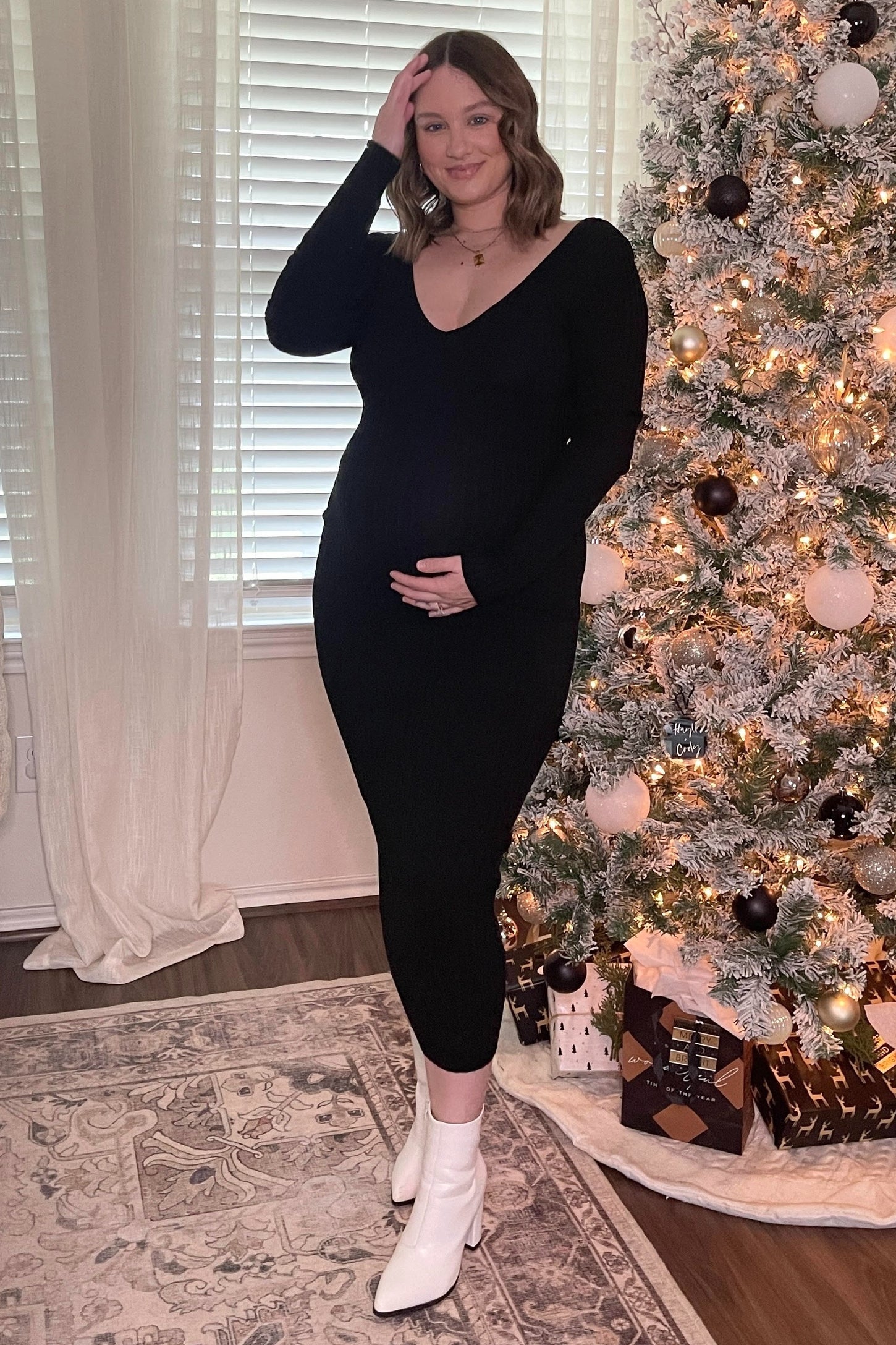 woman wearing a black ribbed maternity dress and holding her baby bump in front of a decorated Christmas tree