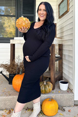 woman wearing a black ribbed maternity dress and holding a pumpkin, showing her baby bump amid autumn decorations