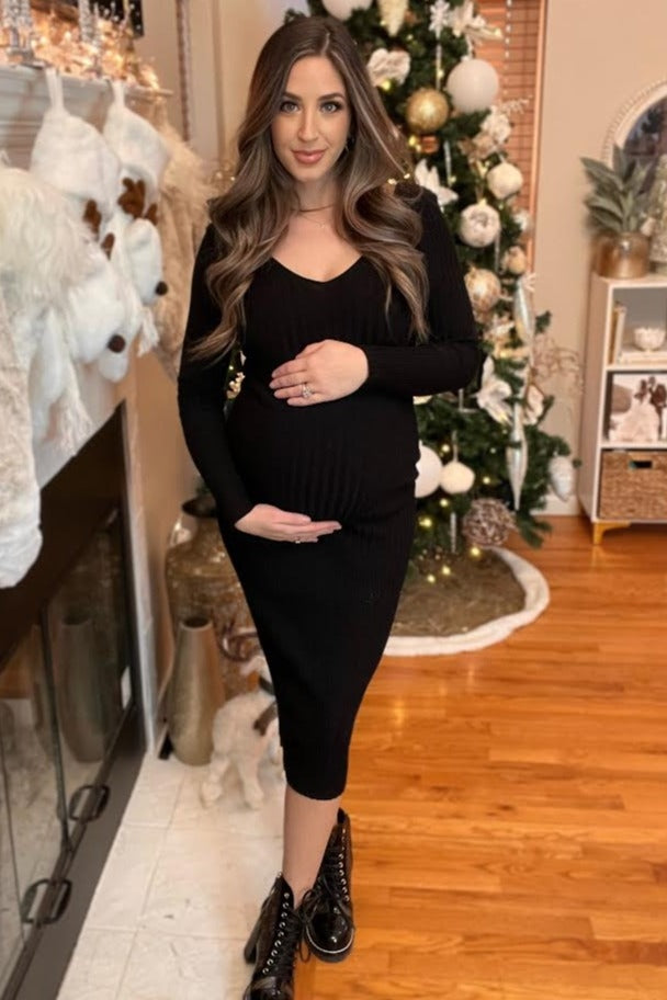 woman wearing a black ribbed maternity dress, holding her baby bump in front of a decorated Christmas tree