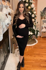 woman wearing a black ribbed maternity dress, holding her baby bump in front of a decorated Christmas tree