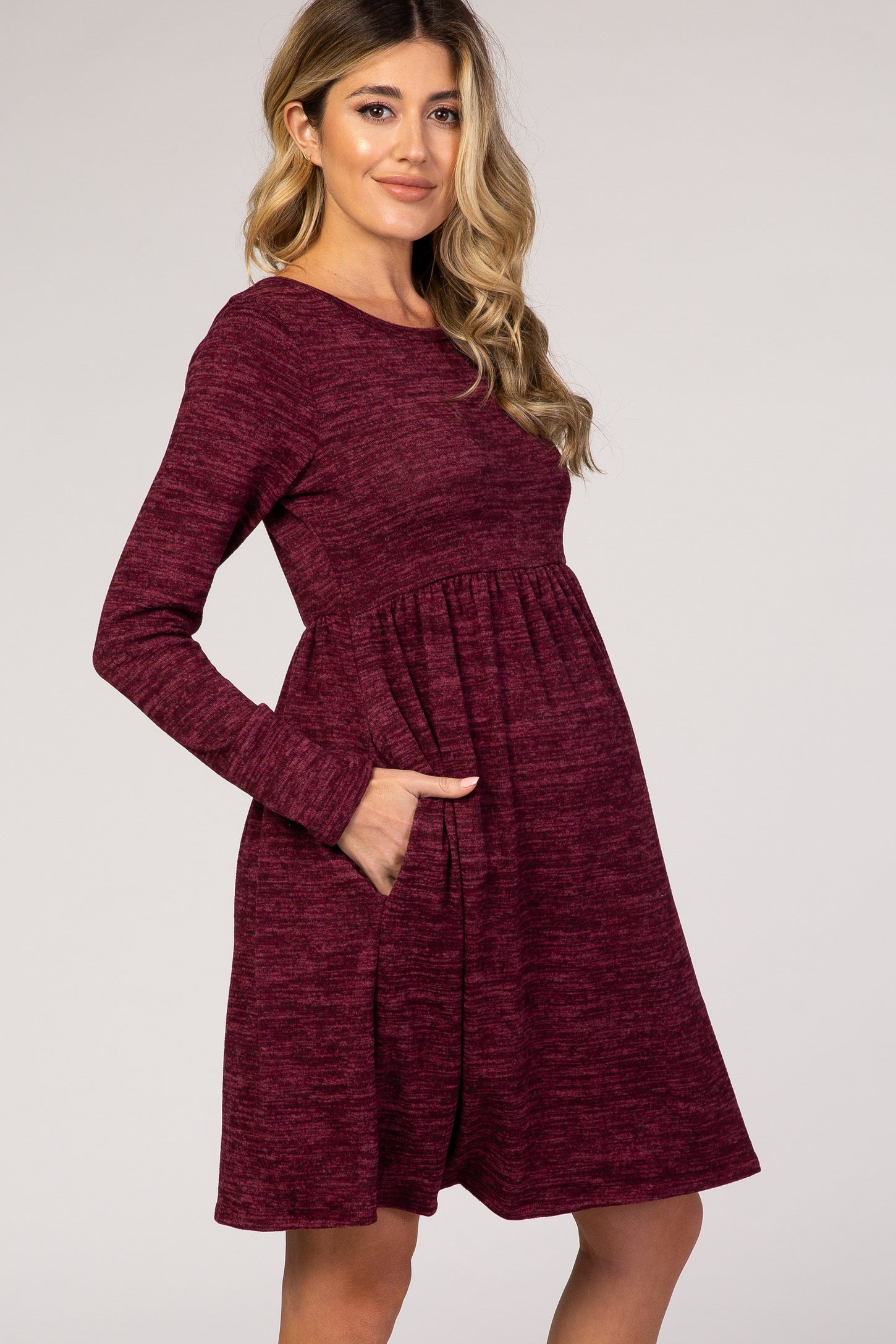 woman wearing a burgundy long sleeve maternity dress with a gathered waist and pockets, showing her baby bump