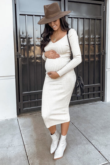 woman wearing a cream ribbed long sleeve maternity dress, holding her baby bump and wearing a brown hat and ankle boots