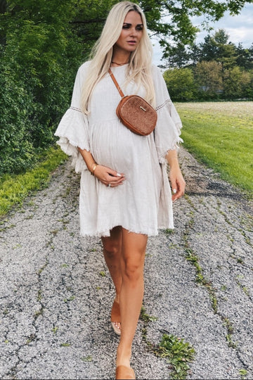 woman wearing a light beige maternity dress with bell sleeves and a baby bump, accessorized with a brown crossbody bag