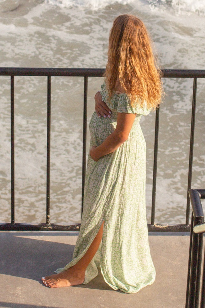 pregnant woman in floral maxi dress holding her baby bump while standing on a balcony overlooking the ocean