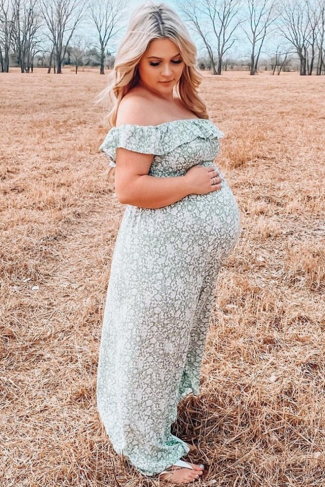woman wearing a light green floral off-the-shoulder maternity dress with a visible baby bump in a field