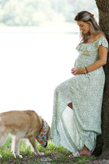woman wearing a green floral maternity dress with off-the-shoulder sleeves and holding her baby bump while standing by a dog