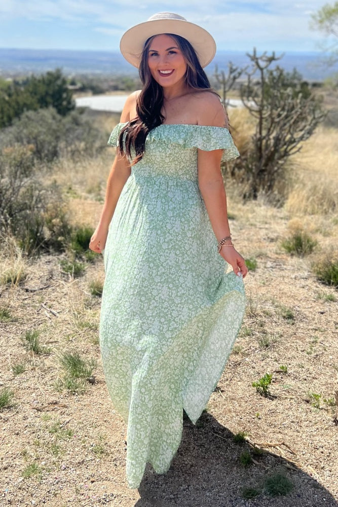 woman wearing a light green off-the-shoulder floral maxi dress and a wide-brimmed hat in a natural outdoor setting