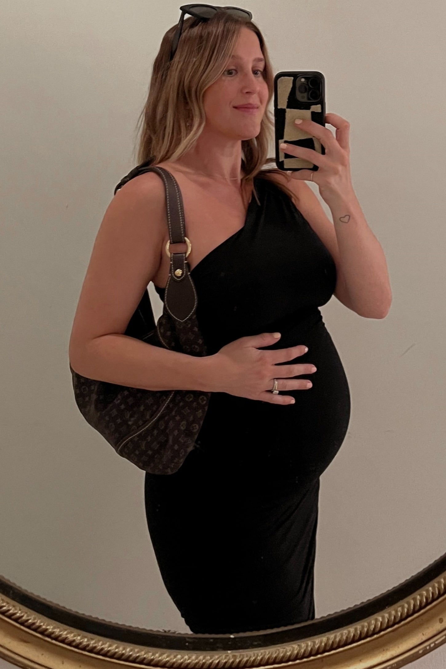 woman wearing a black one-shoulder maternity dress and holding her baby bump while posing in front of a mirror