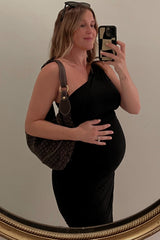 woman wearing a black one-shoulder maternity dress and holding her baby bump while posing in front of a mirror