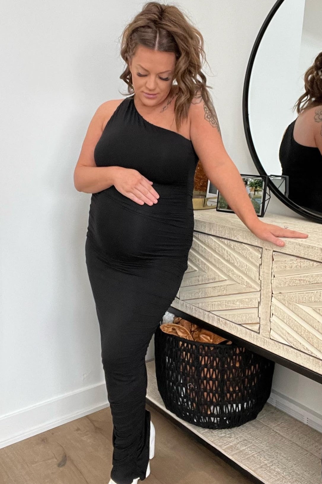 woman wearing a black one-shoulder maternity dress and gently cradling her baby bump while standing by a dresser