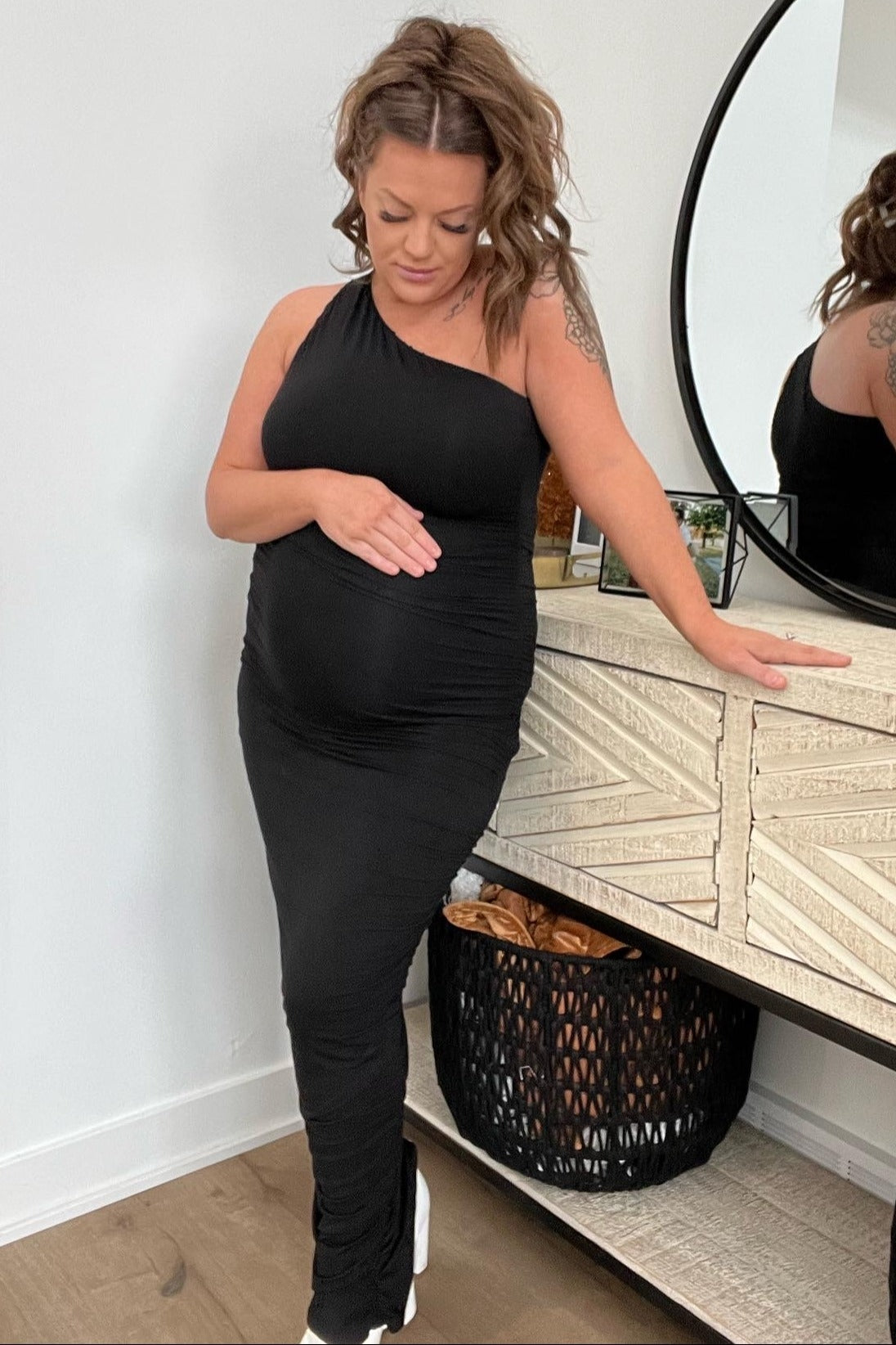 woman wearing a black one-shoulder maternity dress and cradling her baby bump while standing next to a dresser