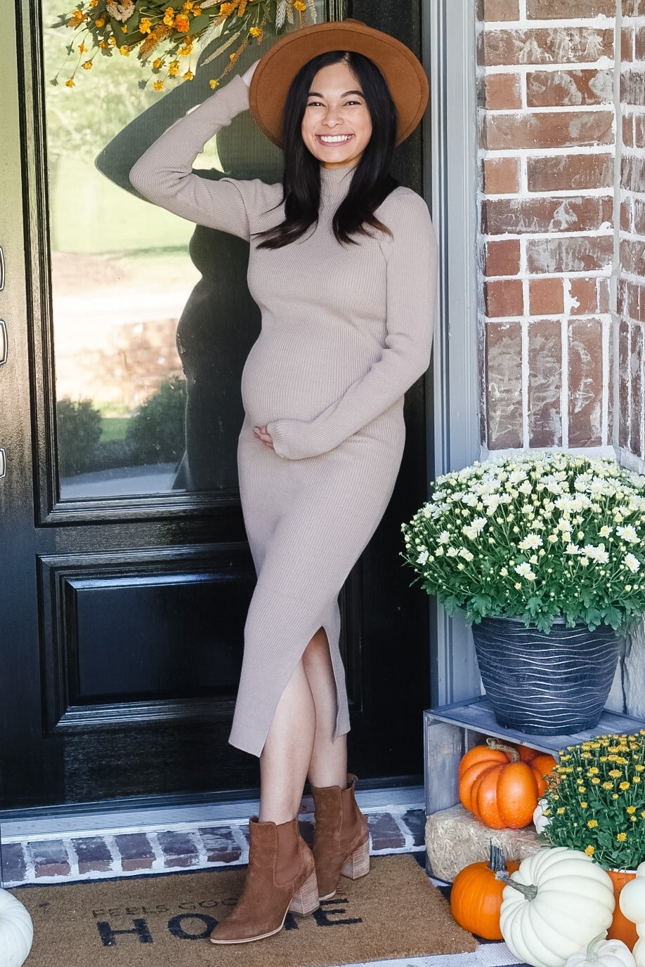 woman wearing a beige ribbed maternity dress with side slits and a brown hat, holding her baby bump at the front door